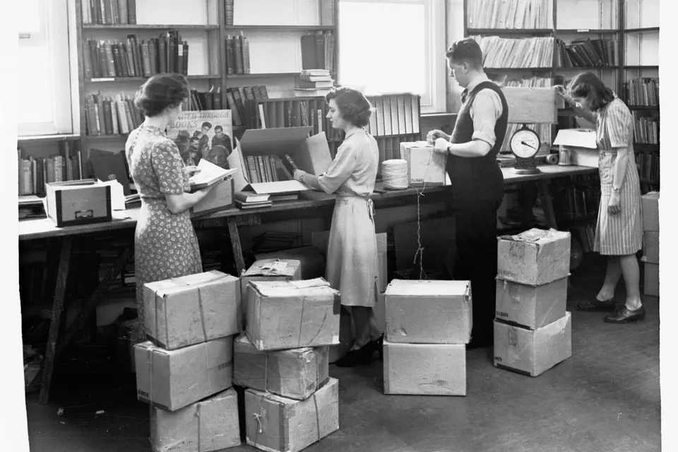 Four people pack cardboard boxes of books in a library, with one weighing a parcel on a scale and another tying string around a box.