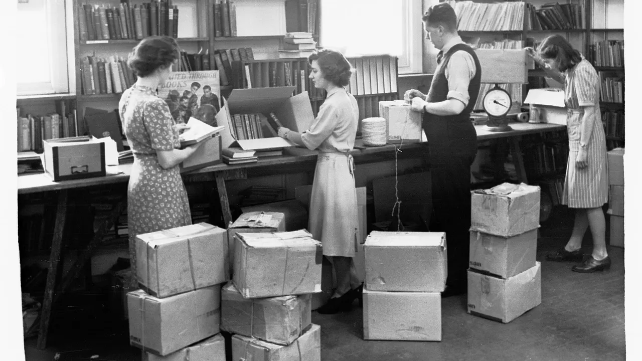 Four people pack cardboard boxes of books in a library, with one weighing a parcel on a scale and another tying string around a box.