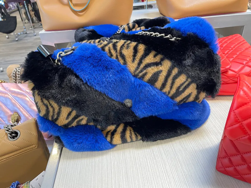 A blue, black and tiger-stripe faux fur handbag with silver chain strap displayed on a white shelf among tan and red bags.