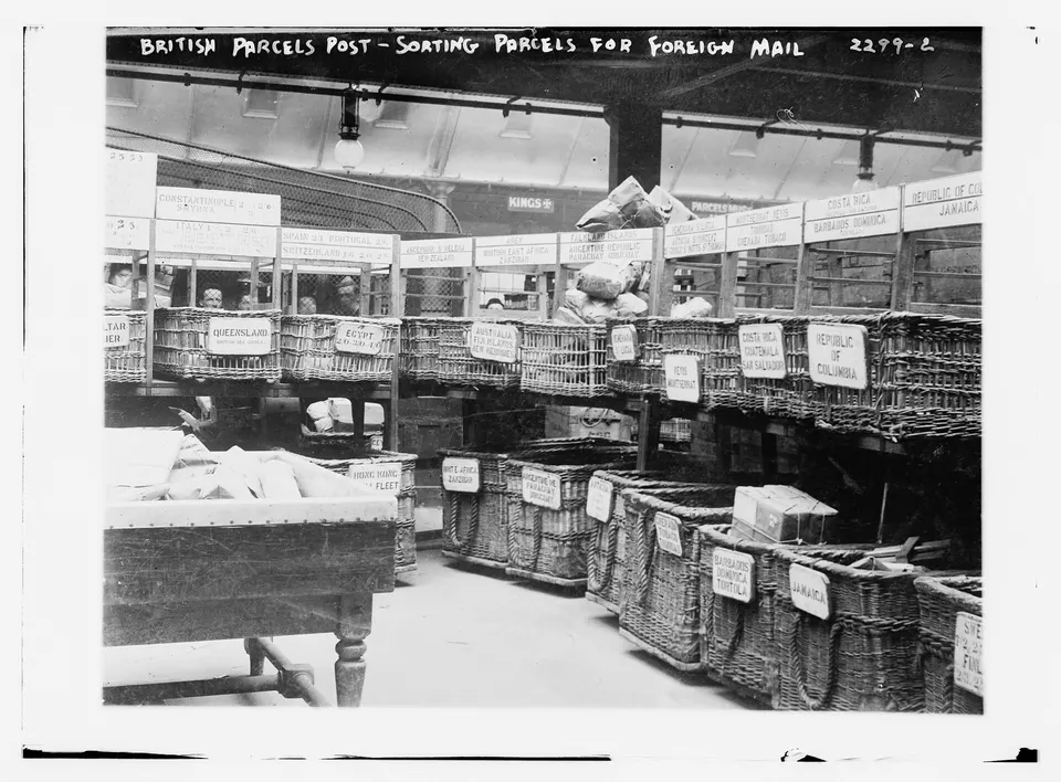 Black-and-white photo of wicker baskets with labels on shelves and tables in a postal sorting room, with parcels and sacks visible.