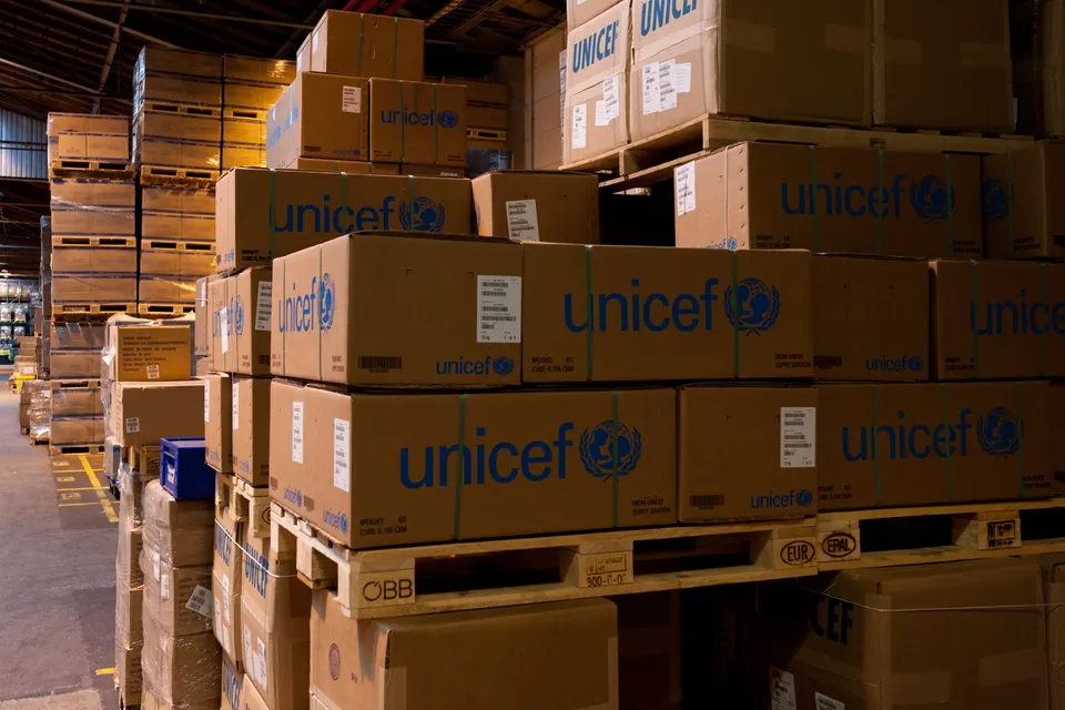Stacks of brown cardboard cartons printed with blue UNICEF logos sit on wooden pallets in a large warehouse with storage racks behind.
