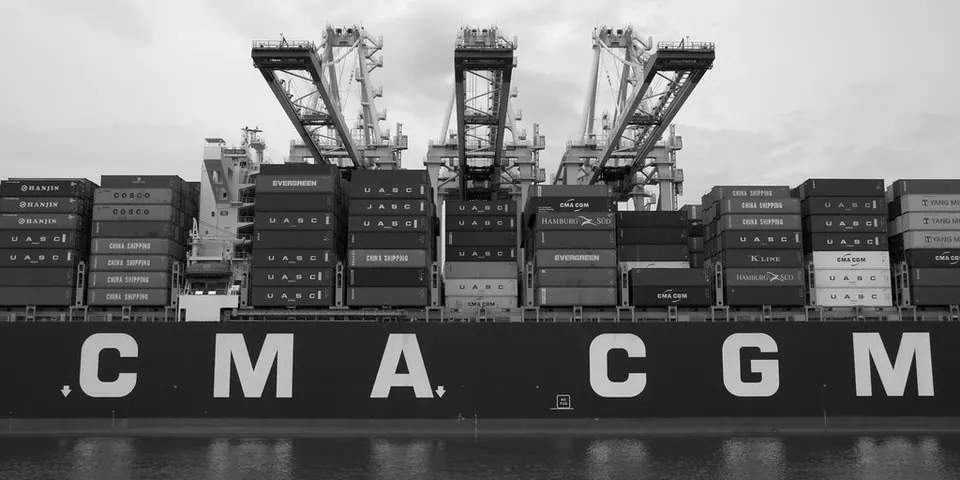 Black and white view of a large container ship with stacked containers and cranes overhead, with CMA CGM lettering on its hull above the water.