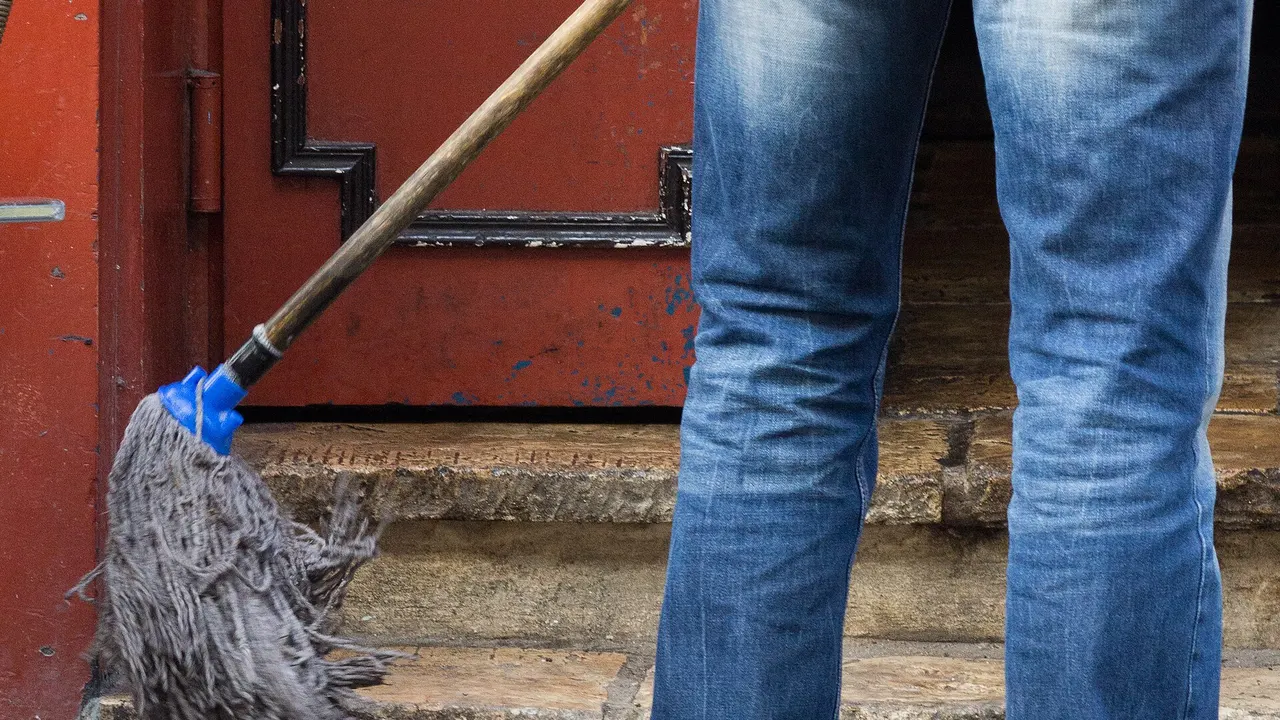 A person in jeans and a dark sweater mops red-painted steps with a grey mop and blue handle, next to a light blue bucket.