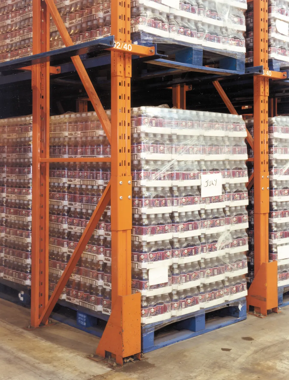 Orange pallet racking in a warehouse holding shrink-wrapped pallets of plastic bottled drinks, with a handwritten 'JULY' label on one pallet.