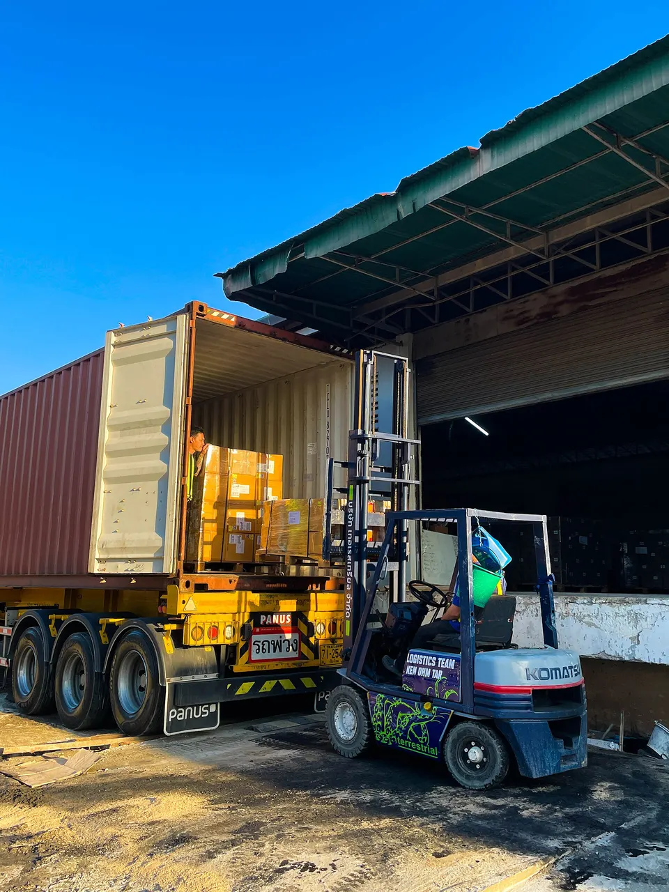 A forklift loads cardboard boxes on pallets from a warehouse into an open shipping container on a yellow truck trailer, under a clear blue sky.