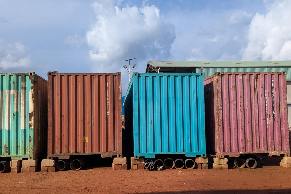 Row of colorful shipping containers in red, teal, and pink sitting on reddish dirt ground under a partly cloudy blue sky.