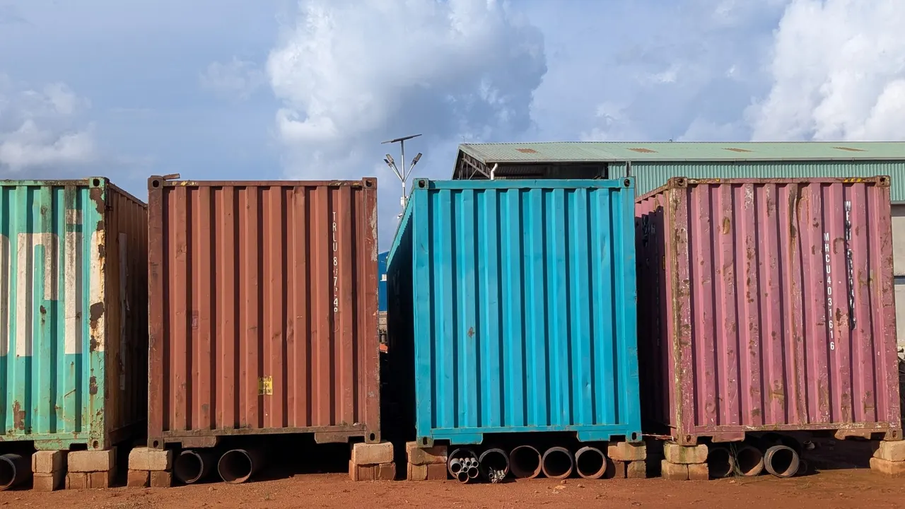 Row of colorful shipping containers in red, teal, and pink sitting on reddish dirt ground under a partly cloudy blue sky.