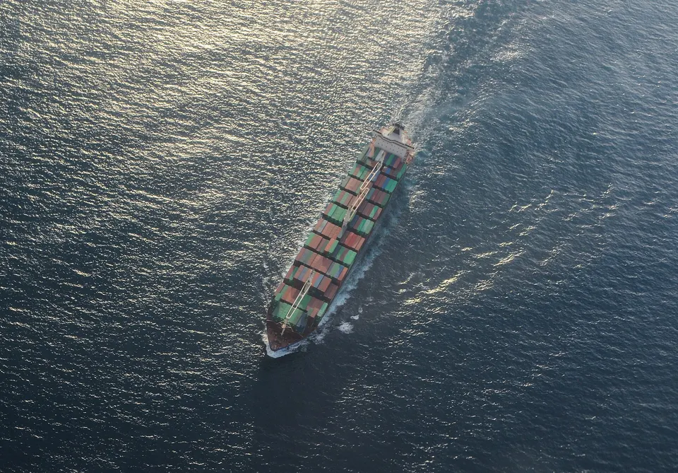 Aerial view of a container ship with stacked green and red containers sailing across dark blue sea with a wake behind it.