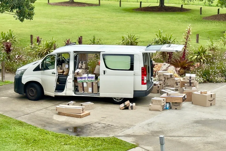 A white delivery van with its side and rear doors open, surrounded by many cardboard parcels on a driveway beside a grassy lawn and tropical plants.