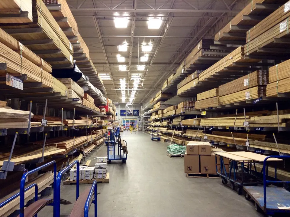 Interior of a large warehouse with lumber stacked on cantilever racks along both sides and blue flatbed carts on the concrete floor.