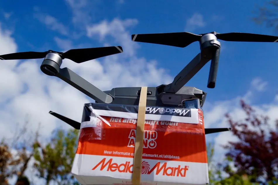 A drone carrying a red and white MediaMarkt parcel box is shown against a blue sky with clouds and trees.