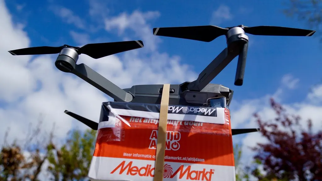 A drone carrying a red and white MediaMarkt parcel box is shown against a blue sky with clouds and trees.