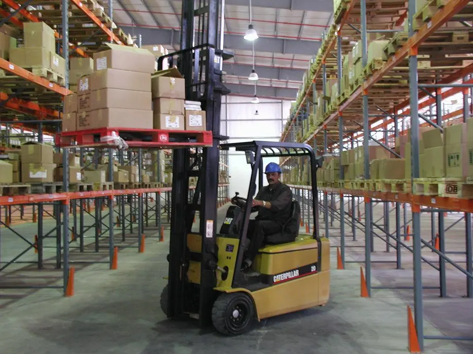 A man in a blue hard hat drives a yellow Caterpillar forklift carrying cardboard boxes on a pallet between tall warehouse pallet racks.