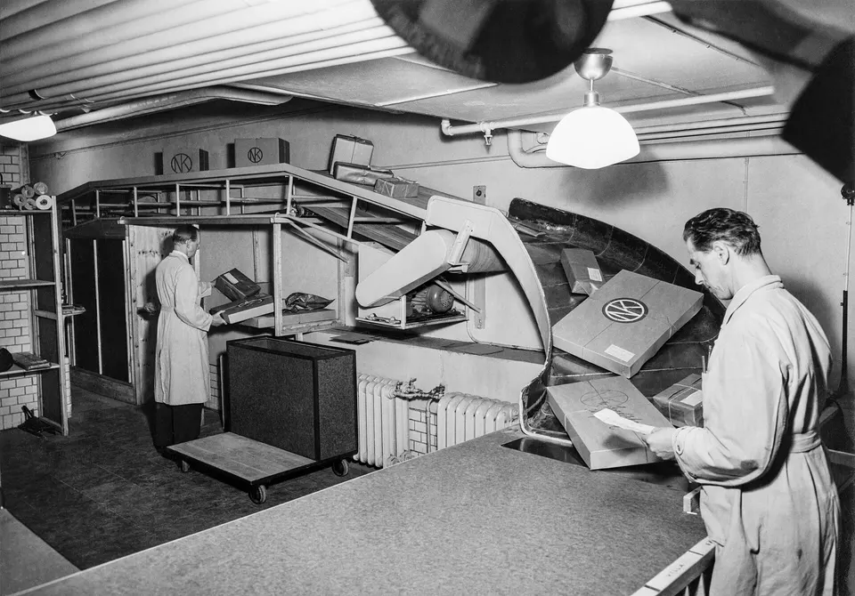 Black-and-white photo of workers in a warehouse-style room handling boxes on a conveyor and cart, with a man in a work coat reviewing a package.