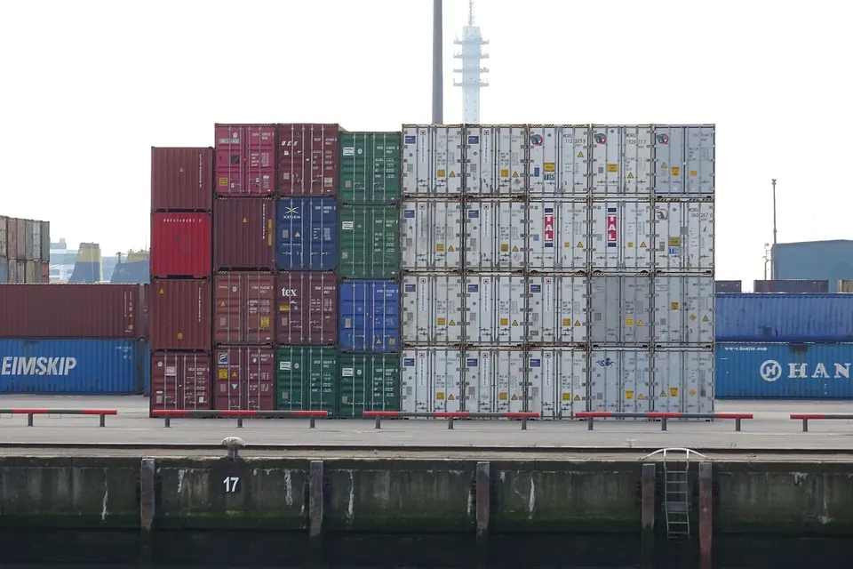 Stacks of shipping containers in red, green, blue, and white stand on a port quay beside dark water, with a tall tower in the background.