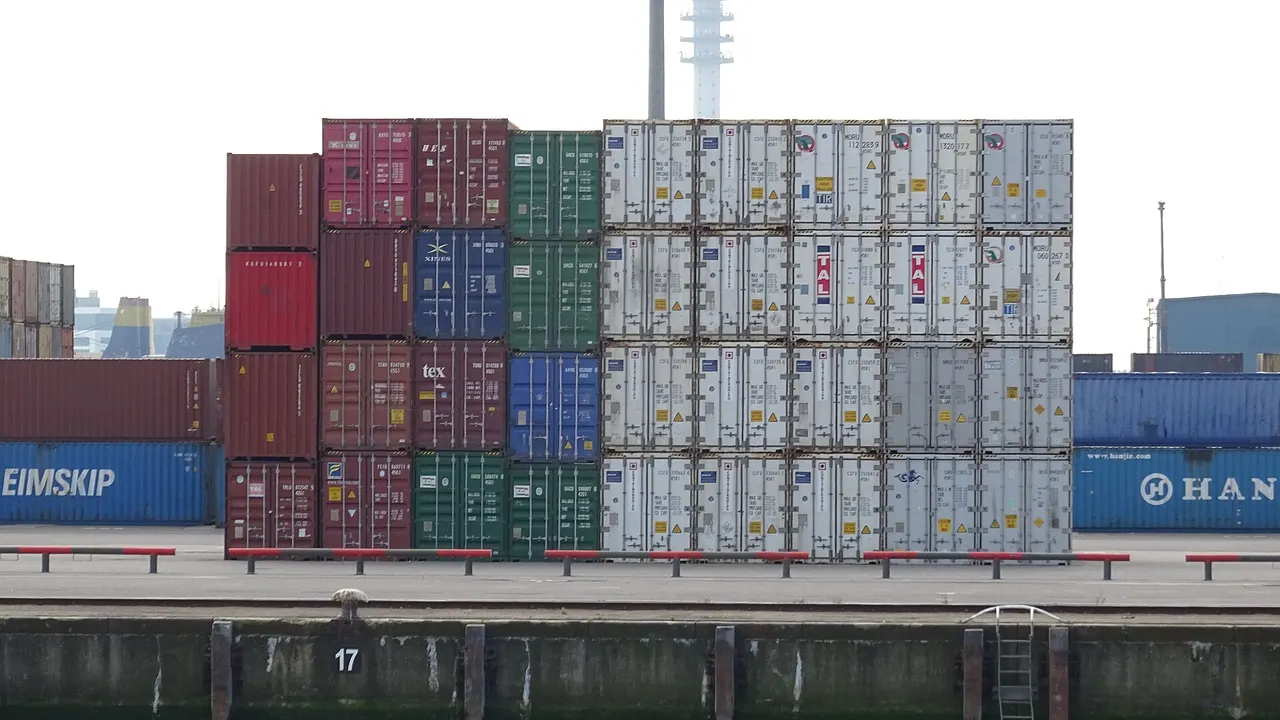 Stacks of shipping containers in red, green, blue, and white stand on a port quay beside dark water, with a tall tower in the background.