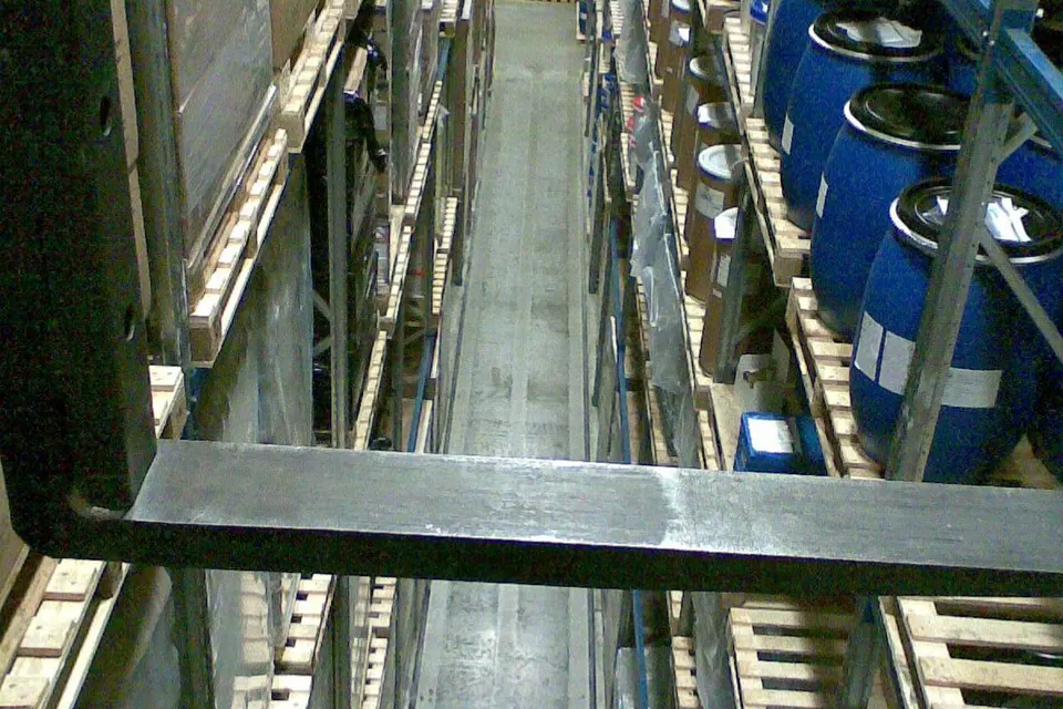 Narrow aisle between tall pallet racks holding stacked cardboard boxes and blue drums, viewed from a forklift's forks in a warehouse.