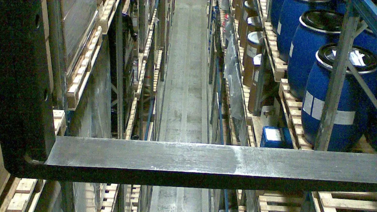 Narrow aisle between tall pallet racks holding stacked cardboard boxes and blue drums, viewed from a forklift's forks in a warehouse.