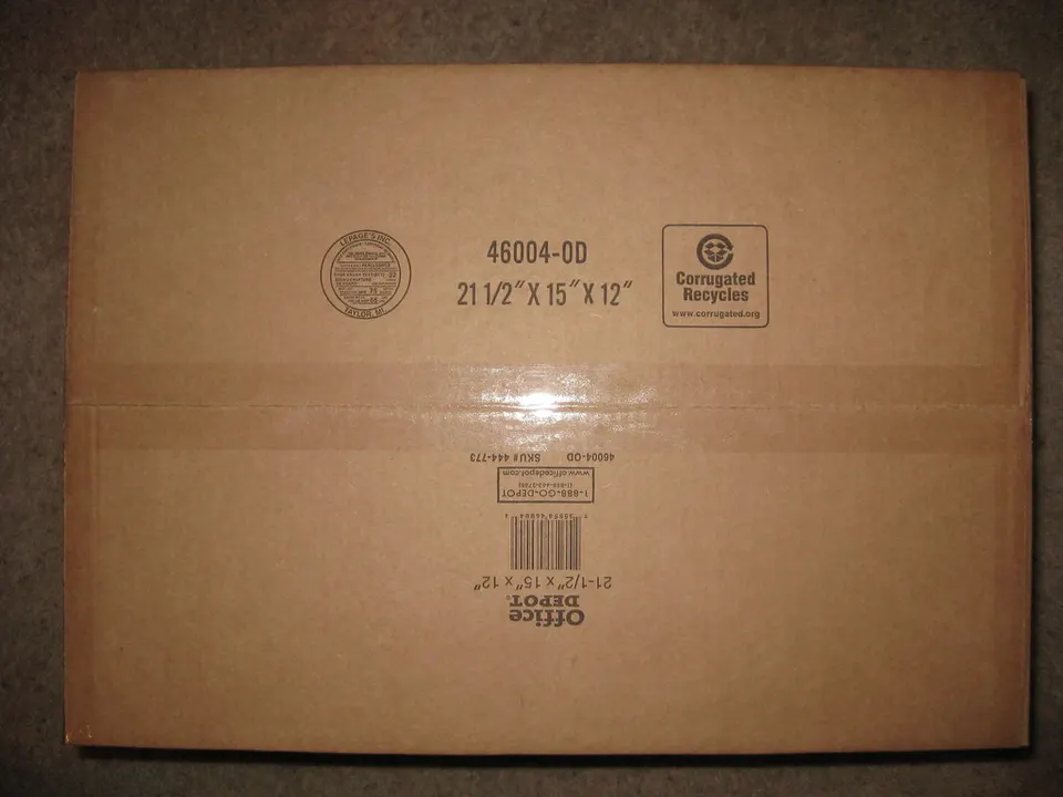 A plain brown corrugated cardboard shipping box with printed size text, a recycling logo, and a barcode label, photographed flat-on against a grey background.