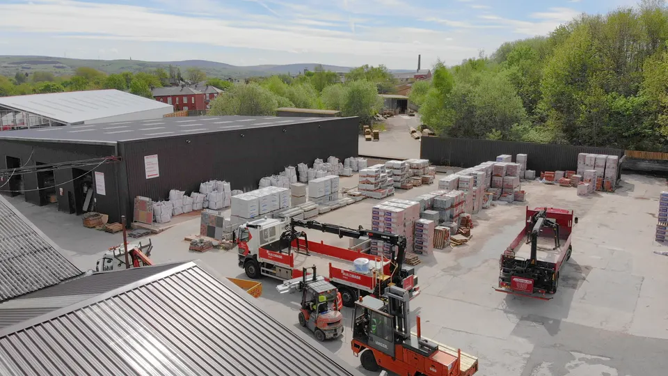 Aerial view of an outdoor yard with stacked pallets of building materials, forklifts, and red trucks with cranes beside a dark warehouse building.