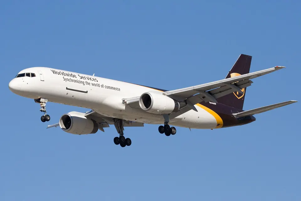 A white cargo jet with purple and gold tail livery and the text 'Worldwide Services' on its fuselage flies against a clear blue sky with landing gear extended.