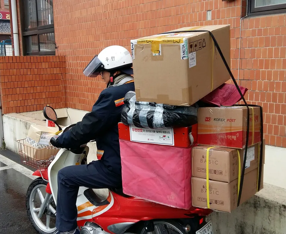 A postal worker in a white helmet and dark uniform rides a red scooter loaded with stacked parcels, including boxes marked Korea Post and Basic, against a brick wall.