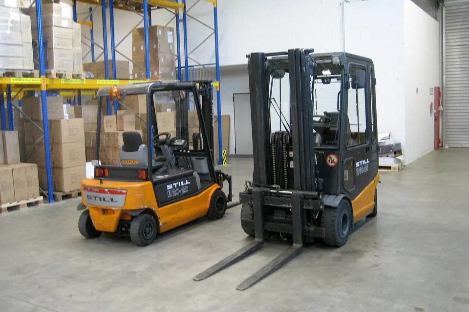 Two forklifts with STILL branding stand in a warehouse aisle beside tall blue racking loaded with cardboard boxes and pallets.
