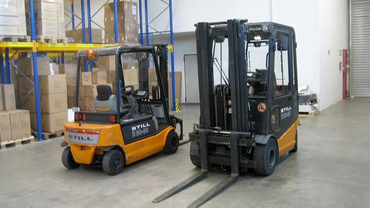 Two forklifts with STILL branding stand in a warehouse aisle beside tall blue racking loaded with cardboard boxes and pallets.
