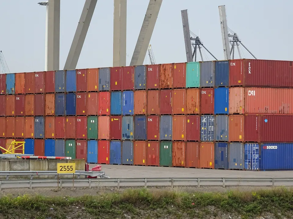 Tall stacks of colorful shipping containers in red, blue, green and orange line a port yard, with cranes visible behind them.