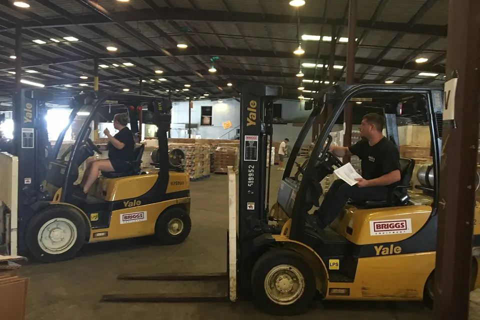 Two workers drive yellow Yale forklifts through a large warehouse with stacked pallets and overhead lights.