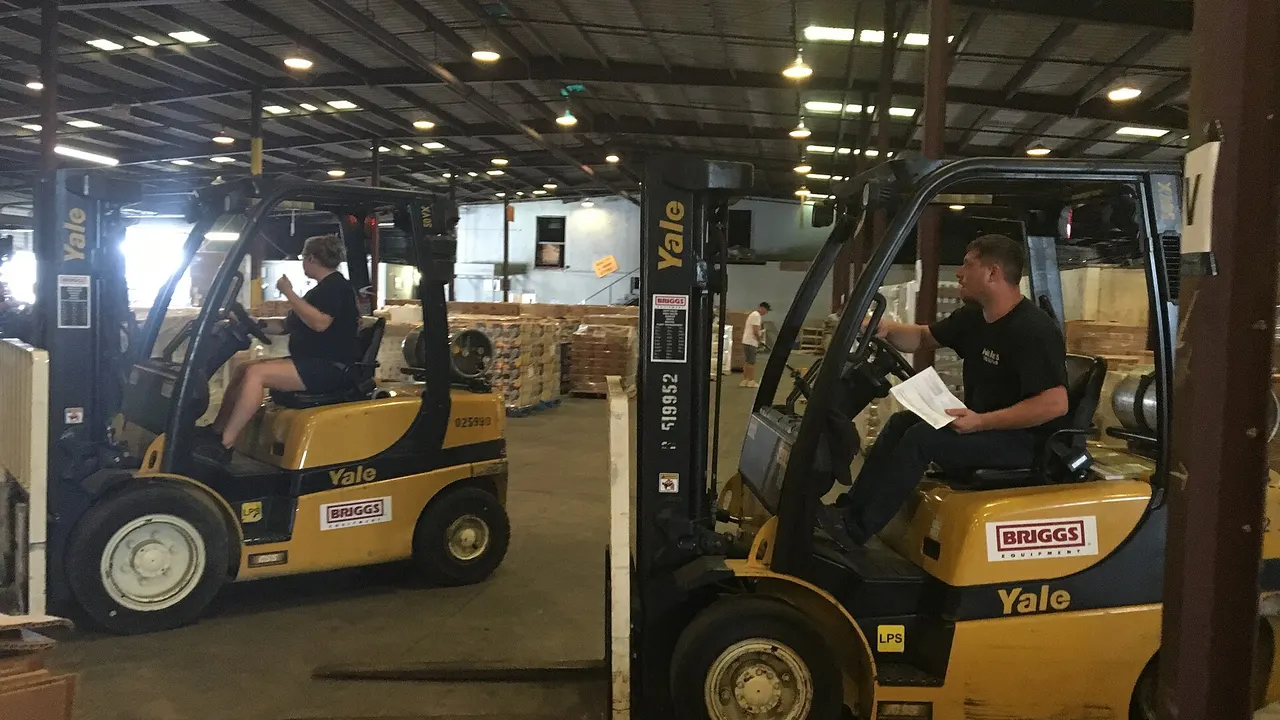 Two workers drive yellow Yale forklifts through a large warehouse with stacked pallets and overhead lights.