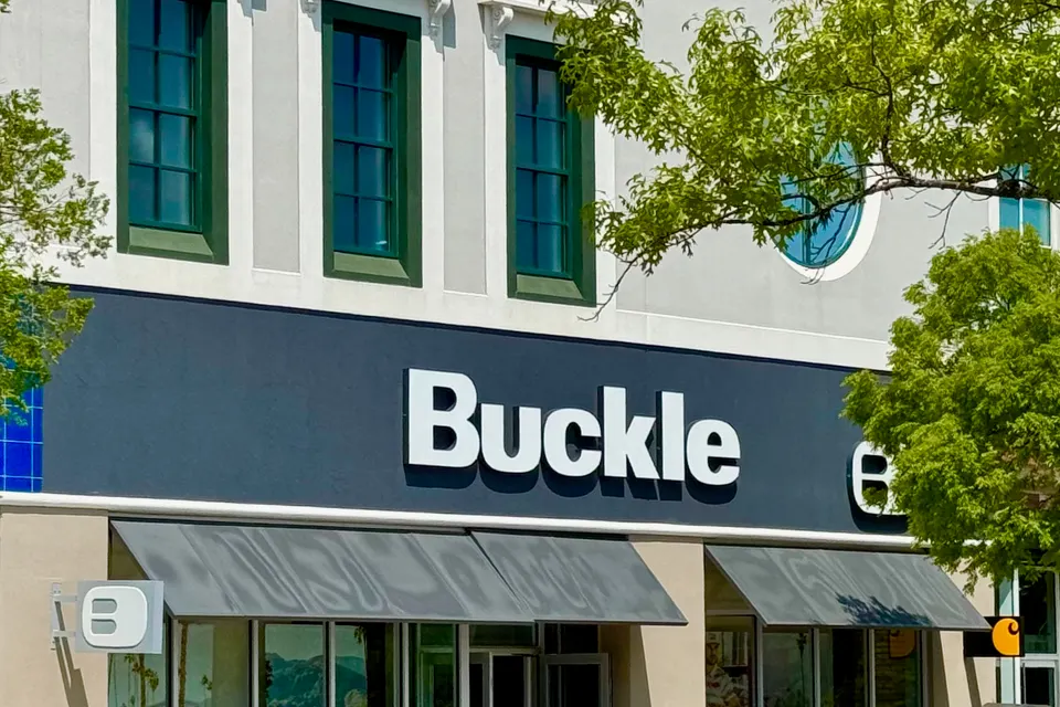 Storefront of a Buckle clothing shop with a dark sign, large white lettering, awnings, and glass windows displaying clothing under a blue sky.