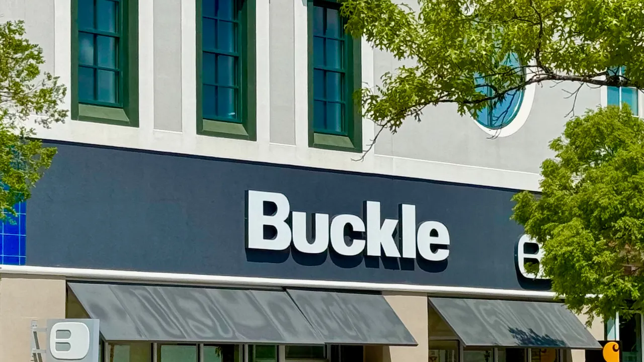 Storefront of a Buckle clothing shop with a dark sign, large white lettering, awnings, and glass windows displaying clothing under a blue sky.