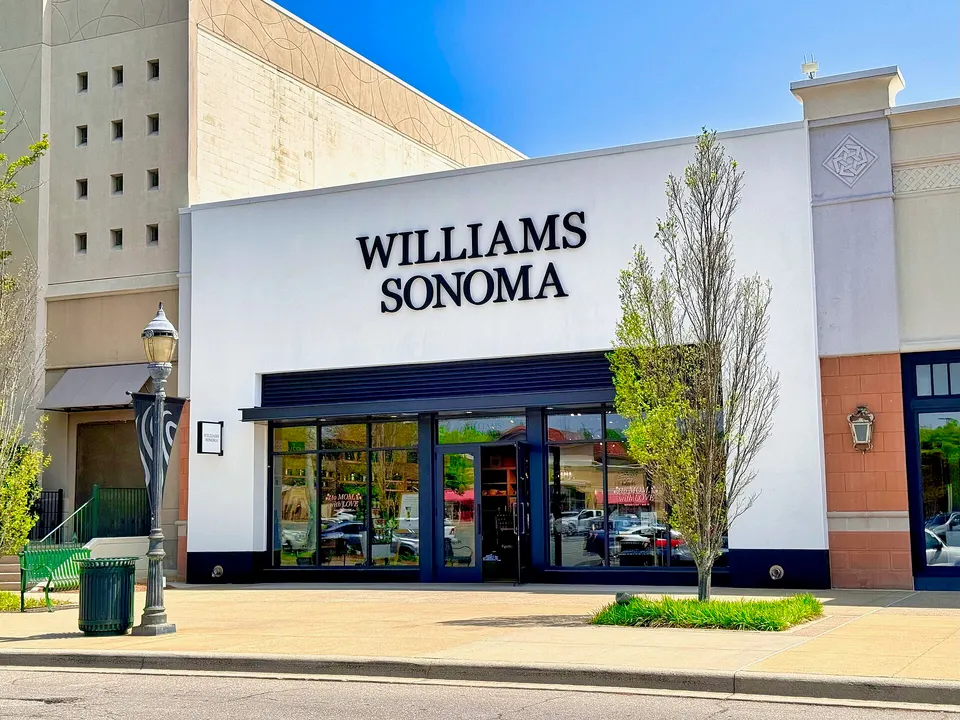 Exterior of a white Williams Sonoma store with black lettering on the facade, a young tree and lamp post on the sidewalk, and a bright blue sky.