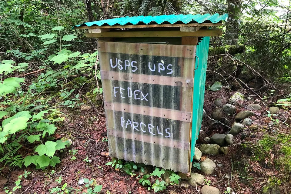 Rustic wooden parcel box in a forest with hand-painted sign reading USPS UPS FEDEX PARCELS, surrounded by rocks and foliage.