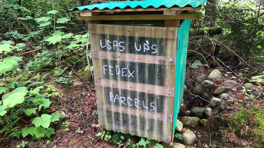 Rustic wooden parcel box in a forest with hand-painted sign reading USPS UPS FEDEX PARCELS, surrounded by rocks and foliage.