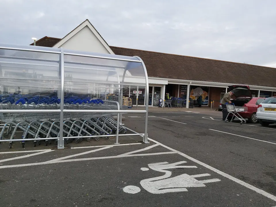 A covered row of shopping trolleys sits outside a retail store, with a shopper loading a trolley beside a red car in the car park.